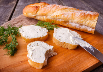 A slice of freshly baked baguette bread and healthy low fat cream cheese with dill and fresh green dill on wooden board.