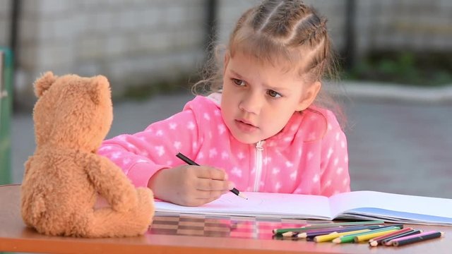 Five-year girl with enthusiasm draws a teddy bear sitting in front of her and a voluntary sticks out his tongue