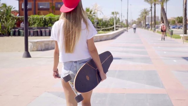 Rear view on single tough young blond female skateboarder in white shirt and backward red hat walking on sidewalk