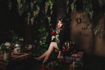 Magic portrait of romantic beautiful girl with wavy hair, red lips, art dress, holding rose flower, sitting on books. Female in scenery of Alice in Wonderland. Fashion fairy tale about princess