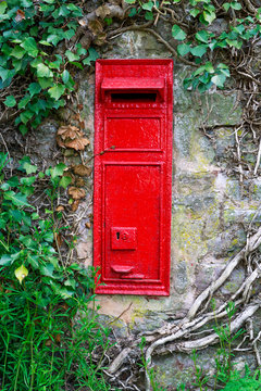 traditional old English red postbox mounted in stone wall surrounded by ivy.