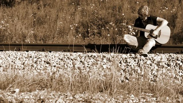 A left-handed long-haired guitarist sitting on the railroad and playing