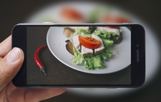 Taking picture of ham sandwich with mobile phone. Phone in male hands.On the plate there is 3 ham sandwiches with tomato and lettuce. Plate is on a black aged wooden table. Vintage style.