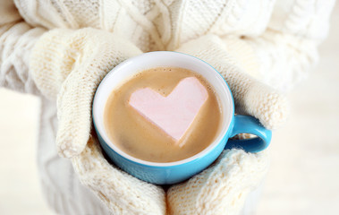Fototapeta na wymiar Female hands in warm mittens holding cup of hot cappuccino with heart marshmallow, close up