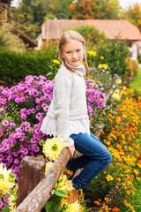 Outdoor portrait of a cute little girl of 8 years old, sitting on a fence, wearing warm grey pullover