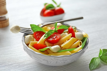 Cooked penne pasta with mozzarella, fresh tomatoes and basil in white bowl on wooden table