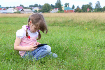 Fototapeta premium Little girl sits and plays with tablet pc at green meadow 