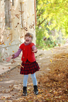 Little beautiful girl in vest throws up leaves in autumn sunny d