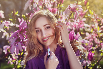 Fototapeta premium Outdoor portrait of a young beautiful happy smiling lady using lipstick near magnolia tree with flowers. Model looking at camera. Girl wearing stylish clothes. Female spring fashion