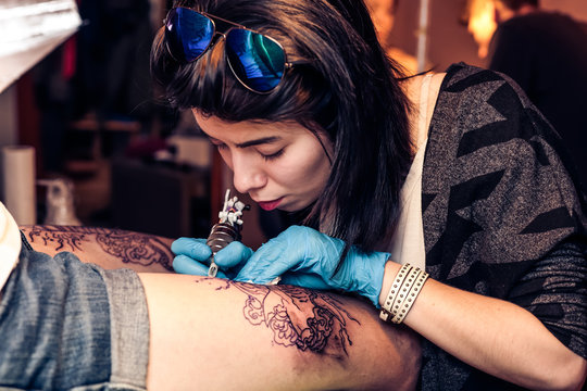 Young beautiful woman tattooer showing process of making a tattoo fill circuit tattoo on leg. Tattoos art on human body skin. Tattooist machine gun.