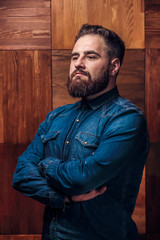 Portrait of Brutal bearded man thinking on a wooden texture in barbershop. Side view of handsome young bearded man keeping arms crossed.