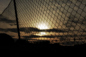 A fence covering the orange-colored sunset sky. Barbed wire on top. Symbolic shot: depression, jail, war.