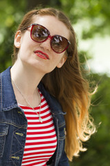 Eine hübsche junge Frau mit langen braunen Haaren im Sommer mit einem rot-weissen Kleid, einer blauen Jeansjacke und einer roten Sonnenbrille.