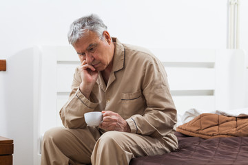 Worried senior man can not sleep. He is sitting on bed and drinking tea.