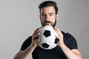Friendly confident soccer player holding ball looking at camera over gray studio background....