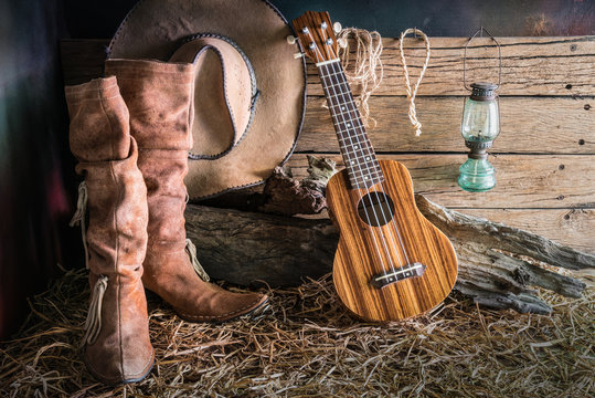 Still life painting photography with ukulele on american west rodeo brown felt cowboy hat and traditional leather boots in vintage ranch barn background