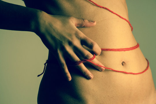 Ventre di giovane donna leggermente di profilo, con mano destra poggiata tra fianco ed ombelico, a tenere un filo rosso intrecciato alle dita ed attorcigliato intorno al corpo.