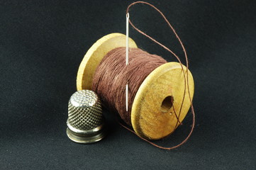 Wooden spool of thread with a needle and a thimble on a dark fabric.