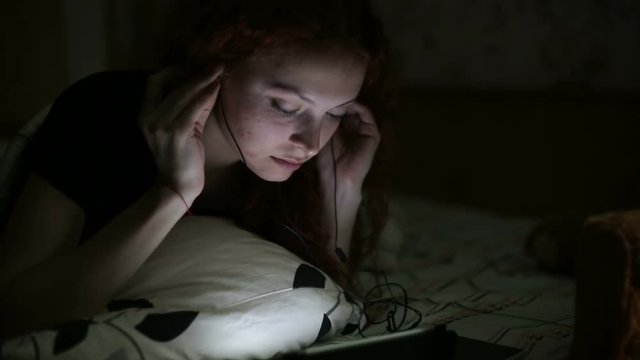 Young woman listening to music with tablet lying on the bed at home in the night