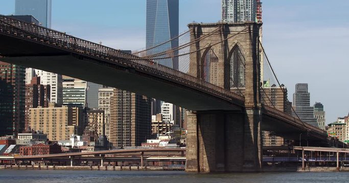 The Brooklyn Bridge stands against a blue sky with the Freedom Tower and other Manhattan skyscrapers in the background while cars criss cross perpetually along the FDR drive