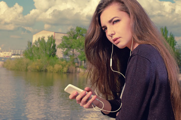 Young woman waiting your call. Dressing in a black wear,  a young caucasian lady is standing by river, holding a mobile phone, with headphones in her ears listening to the music, lost in thought.