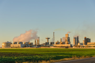 Fototapeta na wymiar Chemical plant with smoke in sunset light on a green field against clear sky