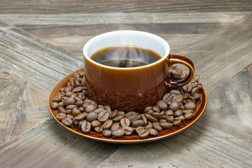 Black hot coffee in brown shiny vintage cup with roasted beans in plate on rustic wooden table.