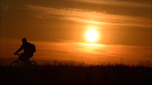  A man riding bicycle on the background of an orange sunset sky, slow motion. Cycling silhouette.  Silhouette of man bicyclist against sunset.
