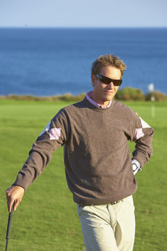 Golfer wearing sunglasses leaning against golf club, hand on hip, looking at camera