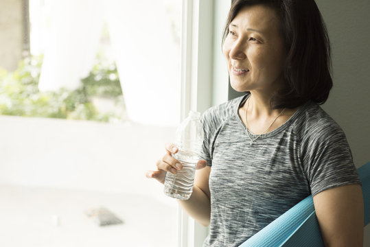 Mature woman holding rolled up yoga mat and bottle of water looking out of window smiling