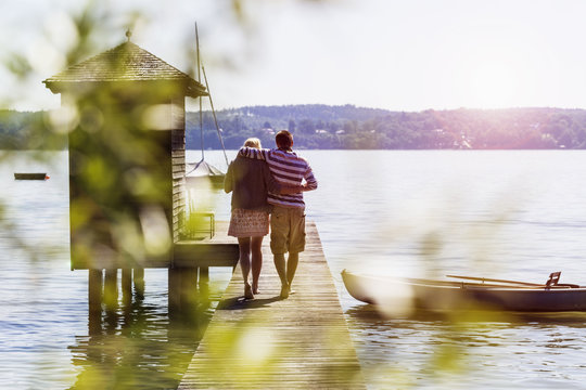 Rear view of young couple walking along pier on lake to boathouse, Schondorf, Ammersee, Bavaria, Germany