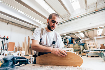 Young man in carpentry workshop looking down sanding skateboard