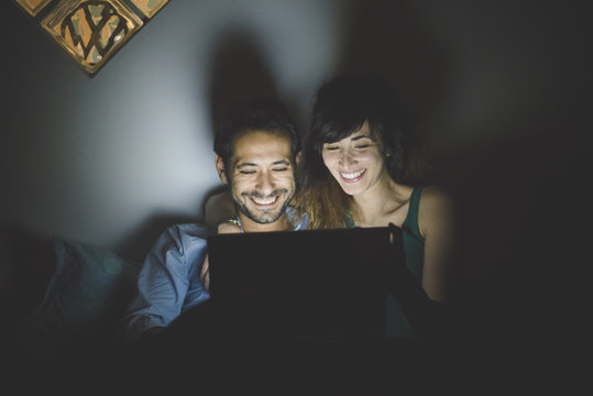 Front view of young couple illuminated by laptop computer screen, looking down smiling