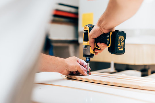 Cropped view of young man in carpentry workshop using cordless screwdriver, screwing into plywood