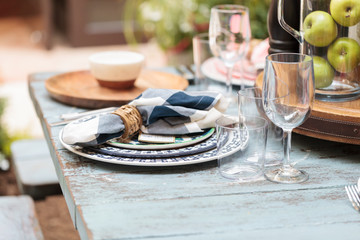 Rustic farm place setting with white and blue plates, napkins and clear wine glasses on a distressed country table on a patio in summer.