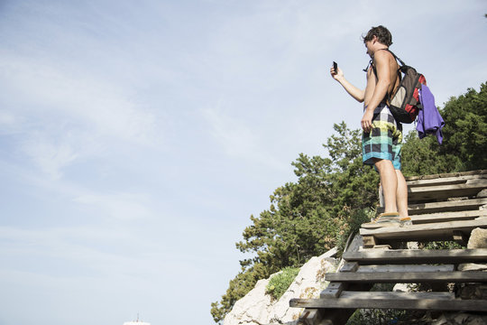 Low angle side view of young man standing on wooden steps using smartphone to take photograph, Cala Luna, Sardinia, Italy