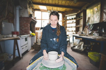 Front view of young woman in workshop sitting at pottery wheel making clay pot, looking down