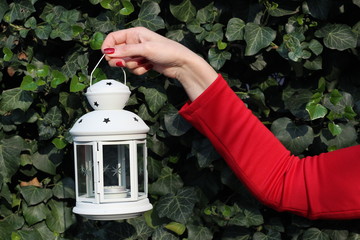 Candle lantern lamp in the hand of girl in red dress on the background of green ivy