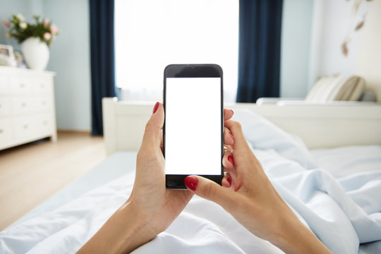Cropped view of female hands holding smart phone with blank screen for your text. Woman reading messages on cell phone with home interior background while resting in bed. Selective focus, film effect
