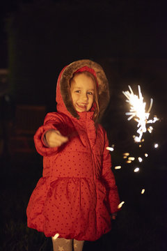 Girl wearing red fur trim coat holding sparkler, looking at camera smiling