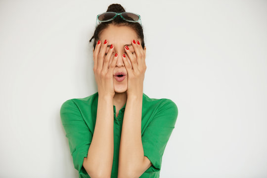 Isolated headshot of funny girl in casual shirt covering her face with both hands. Young female model having fun while posing on white copy space background. Human face expression, body language