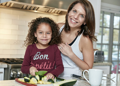 Mother helping daughter chop vegetables in kitchen, looking at camera smiling