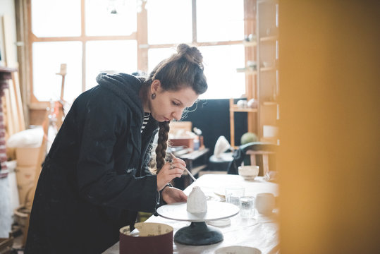 Side view of young woman in workshop applying ceramic glaze to clay pot, looking down