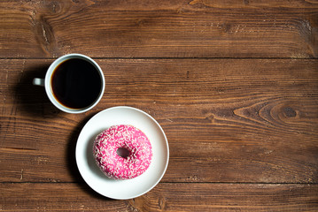 Cup of black coffee with pink donut on wooden background, top view