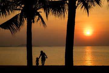 Beautiful sunset on beach with caring happy mother and daughter walking under coconut tree in silhouette twilight golden hour/Sunset on beach, happy mother and daughter