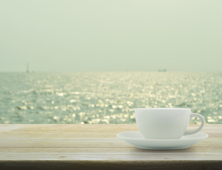 White cup on wooden table with blurred sea waves with bokeh ligh