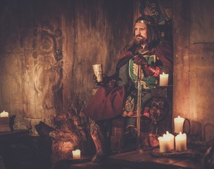Old medieval king with goblet of wine on the throne in ancient castle interior.