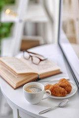 White cup of the coffee and an open book on the table  in a light room..