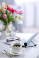 White cup of the coffee and a bouquet of flowers on wooden table  in a light room..