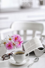 White cup of the coffee and fresh flowers on wooden table in a light room..